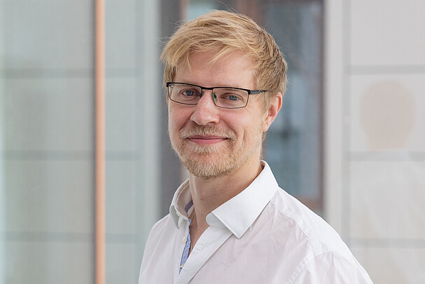 Porträt von Prof. Dr. rer. nat. Henning Wachsmut mit Brille und weißem Hemd, lächelnd vor einem modernen Glas- und Metallhintergrund.