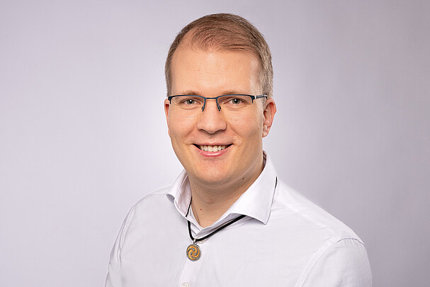 Porträt von Prof. Dr. rer. nat. Marius Lindauer mit Brille, weißem Hemd und Anhänger-Halskette, lächelnd vor einem hellgrauen Hintergrund.